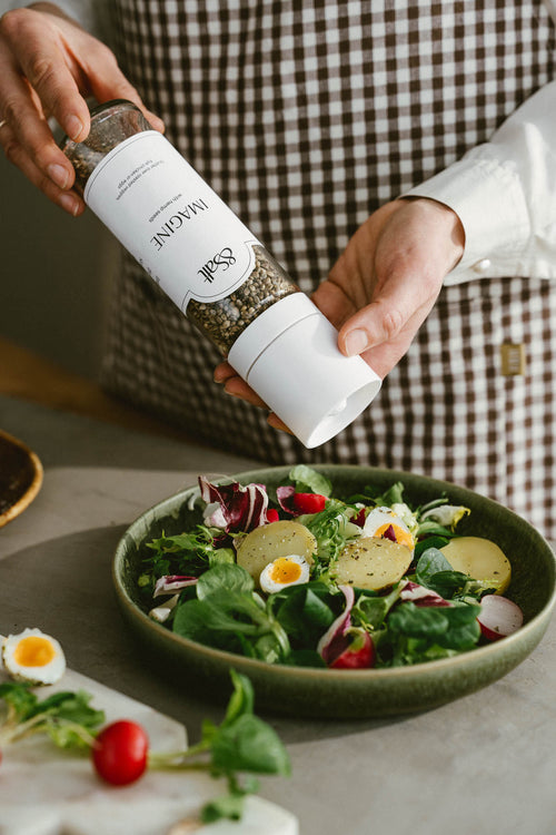 Person seasoning a salad with a bottle of spices, wearing a checkered apron.