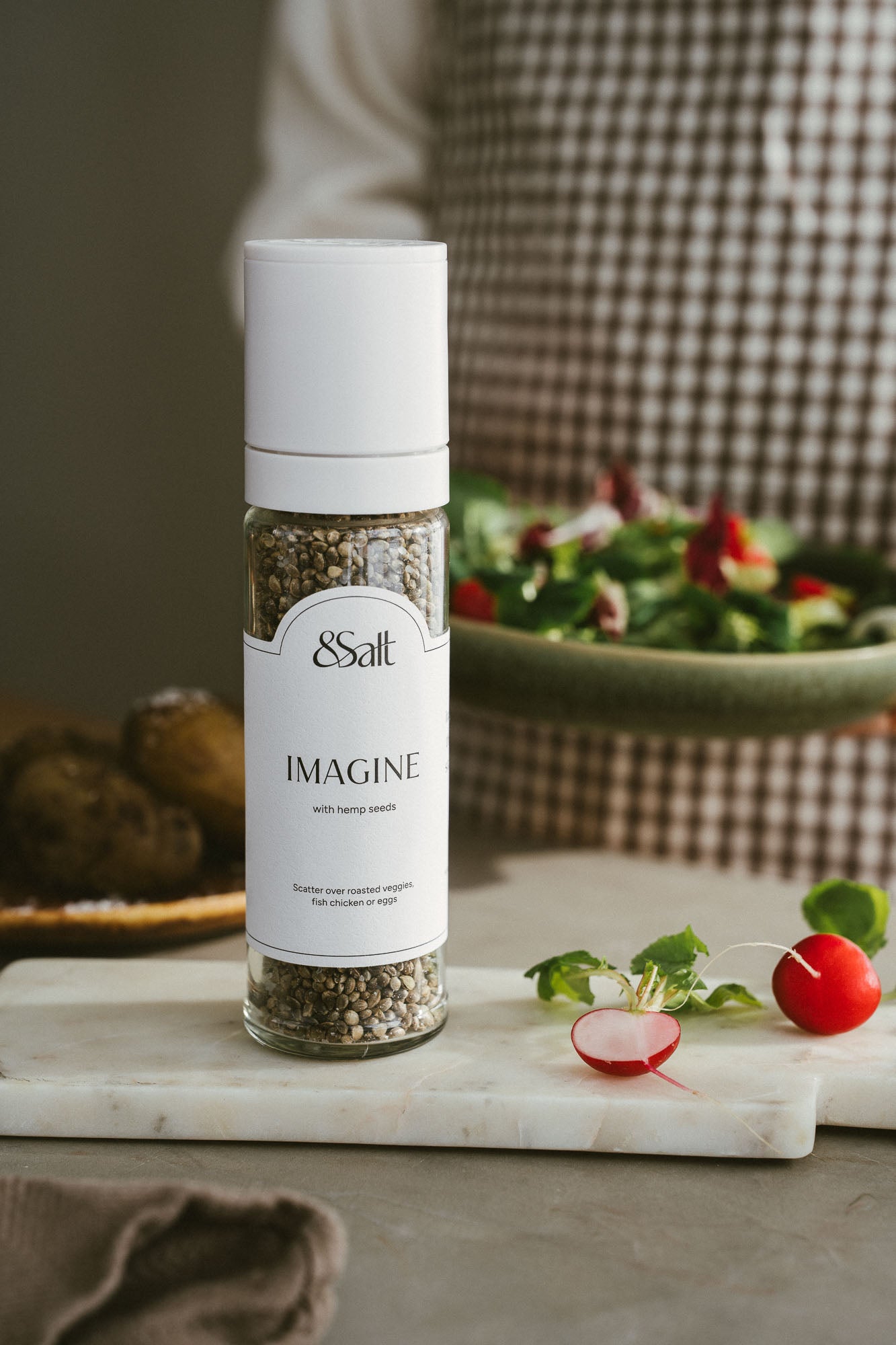 Bottle of '&Salt Imagine' seasoning on a marble cutting board with vegetables and a blurred background.